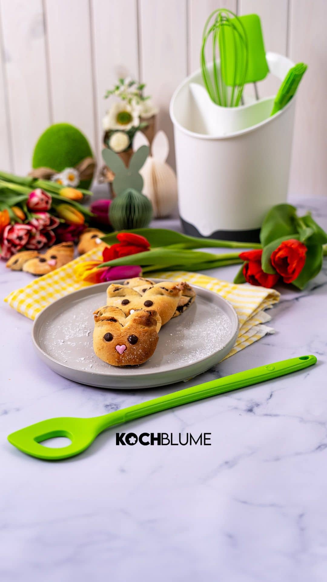 Drei Hasen-förmige Gebäckstücke auf einem Teller, umgeben von Tulpen, Backutensilien und dem KOCHBLUME-Logo.