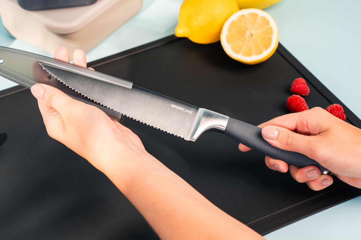 Des mains tenant le « Chefmesser » de Kochblume GmbH, à côté de framboises fraîches, de citrons éclatants et d’une planche à découper noire posée sur un fond bleu vif : un décor idéal pour concocter de délicieuses recettes à base de fruits.