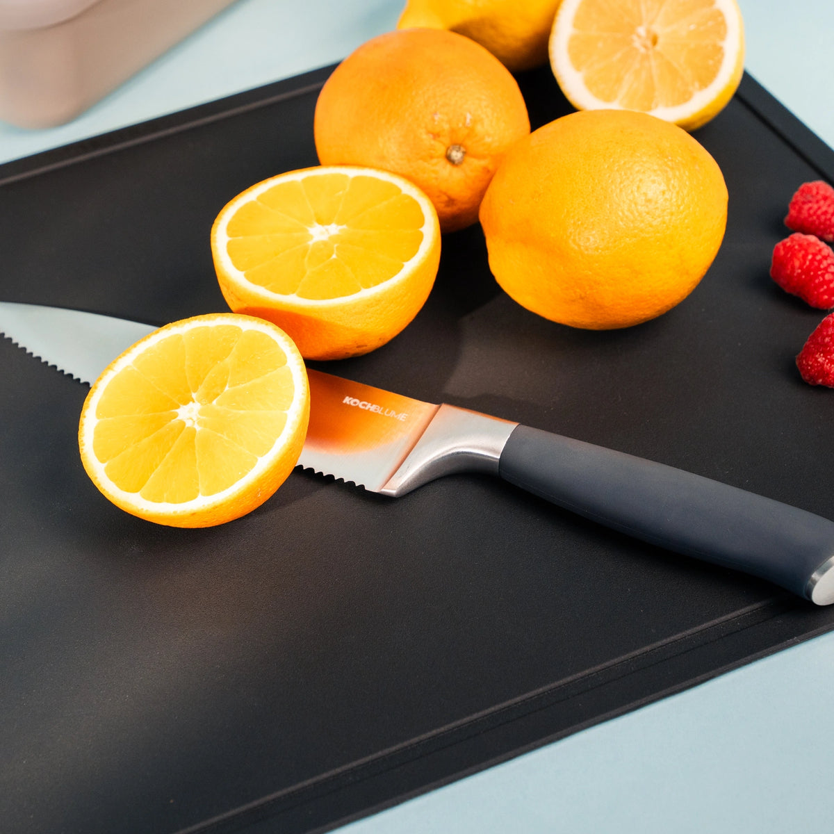 Zu sehen ist ein Schneidbrett „Schneidunterlage TPU“ der Firma Kochblume GmbH, auf dessen schwarzer Oberfläche ein Messer eine Orange schneidet, umgeben von ganzen Orangen und Himbeeren.