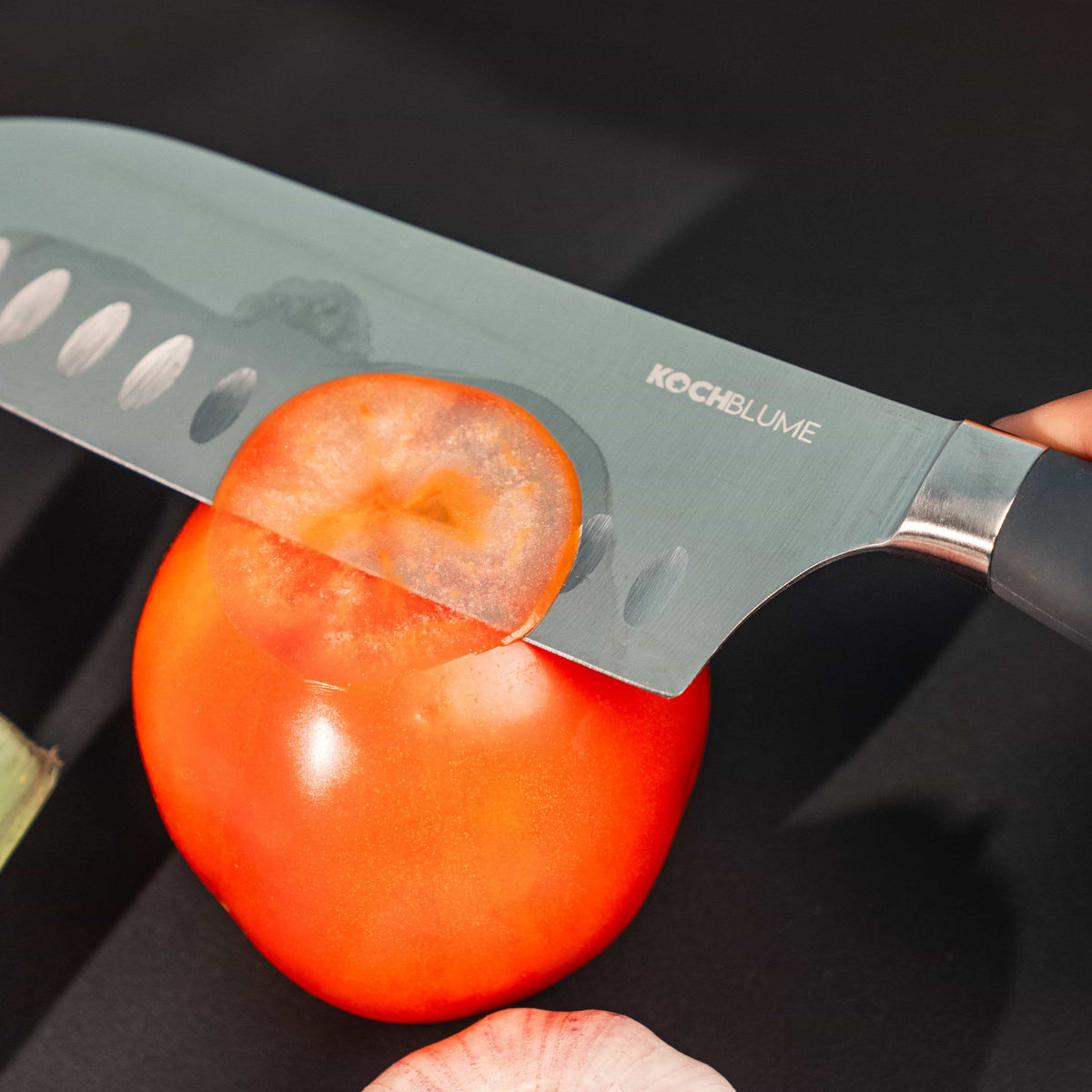 Ein scharfes Santokumesser der Firma Kochblume GmbH schneidet eine reife Tomate auf einer dunklen Unterlage, daneben liegen Knoblauchzehen – eine einfache, aber eindrucksvolle Küchenszene.