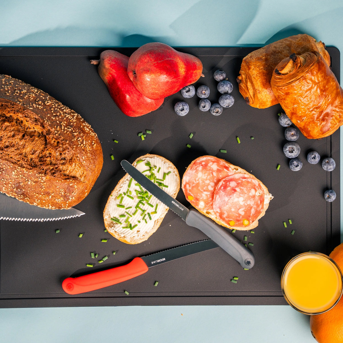 Du pain de mie, du fromage, du salami, des poires, des myrtilles, du jus d'orange et un couteau de petit-déjeuner de la marque Kochblume GmbH, doté d'un manche ergonomique en acier inoxydable, sont disposés sur une planche à découper noire.
