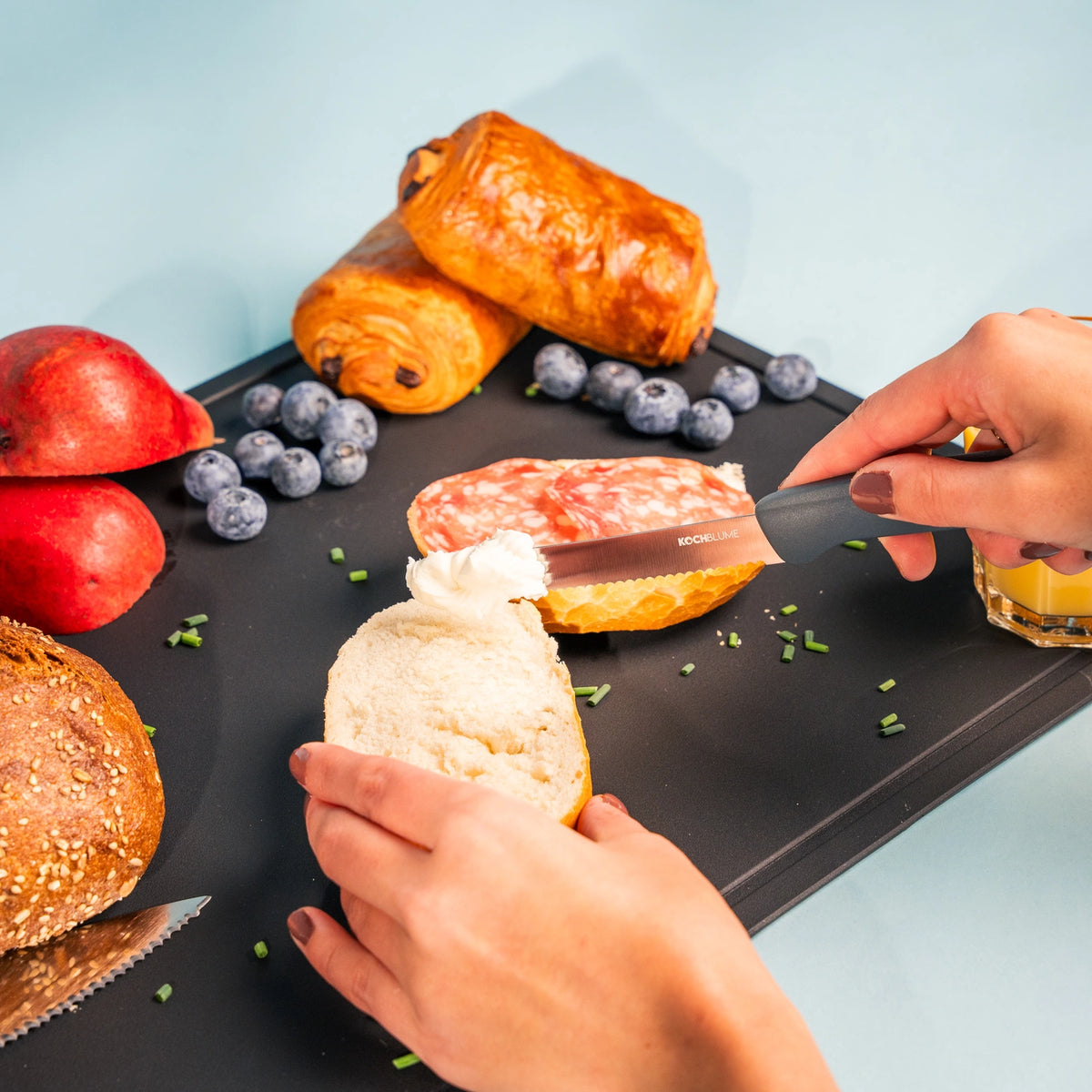 Des mains étalant du fromage à tartiner sur du pain à l'aide du couteau à petit-déjeuner de Kochblume GmbH, doté d'un manche ergonomique et entouré de viennoiseries, de myrtilles, de grenade et d'un verre de jus de fruits. La lame en acier inoxydable garantit un étalement facile et sans effort, pour un petit-déjeuner des plus agréables.