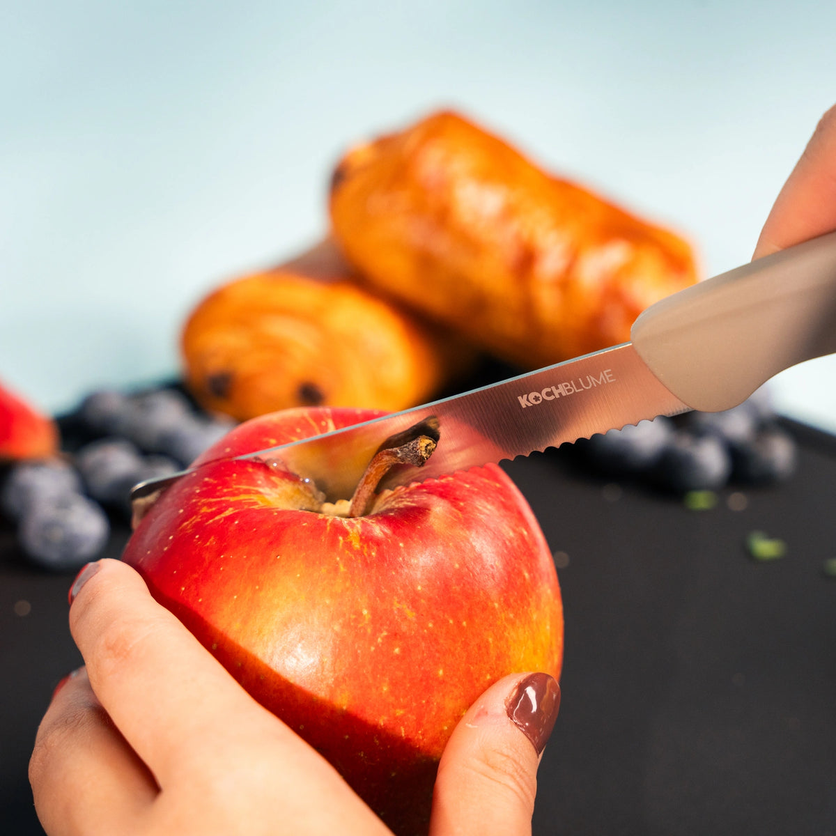 Des mains en train de couper une pomme rouge avec le couteau « Frühstücksmesser » de Kochblume GmbH, doté d'un manche ergonomique ; on aperçoit des pâtisseries et des myrtilles à l'arrière-plan.