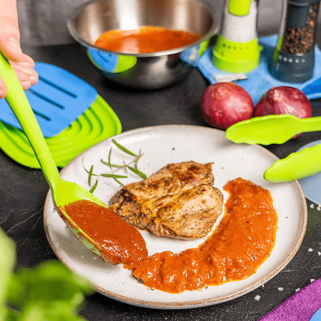 Gegrillte Hähnchenbrust auf einem Teller, daneben wird Tomatensoße auf den Teller gegeben; im Hintergrund sind Küchenutensilien zu sehen.