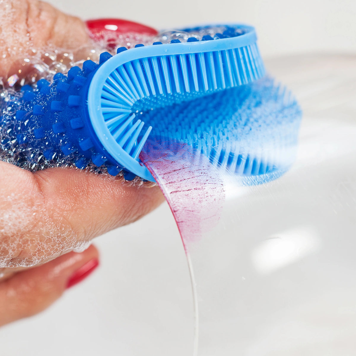 Ein seifiges Glas wird mit dem blauen „Scrubby“ der Kochblume GmbH sauber geschrubbt, der von einer Hand mit rot lackierten Fingernägeln gehalten wird.