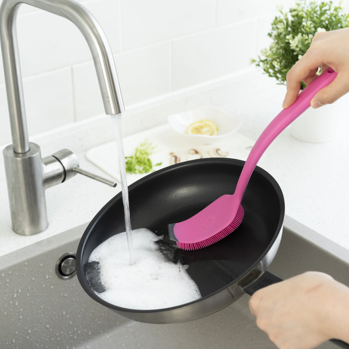 Eine Person schrubbt eine mit Spülmittel eingeseifte Pfanne unter fließendem Wasser aus dem Küchenhahn kräftig mit der rosa Universalbürste der Kochblume GmbH ab und sorgt so dafür, dass alle Rückstände entfernt werden und das Kochgeschirr strahlend sauber wird.