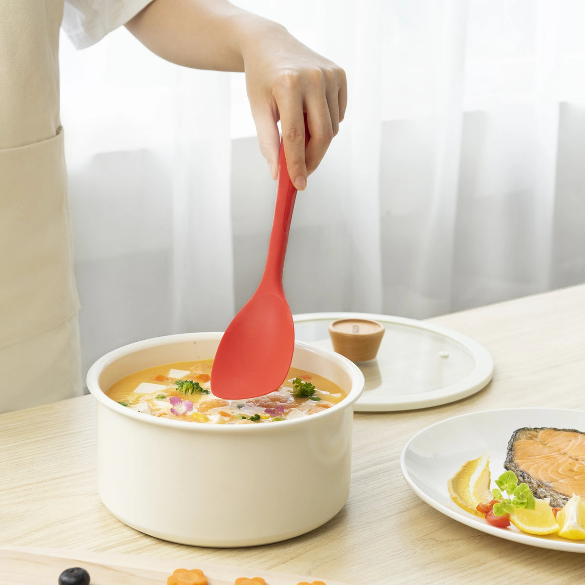 Eine Hand hält den Flexlöffel der Firma Kochblume GmbH und rührt Suppe in einem weißen Topf um; auf einem Teller daneben sind Essensreste und Lachs zu sehen.