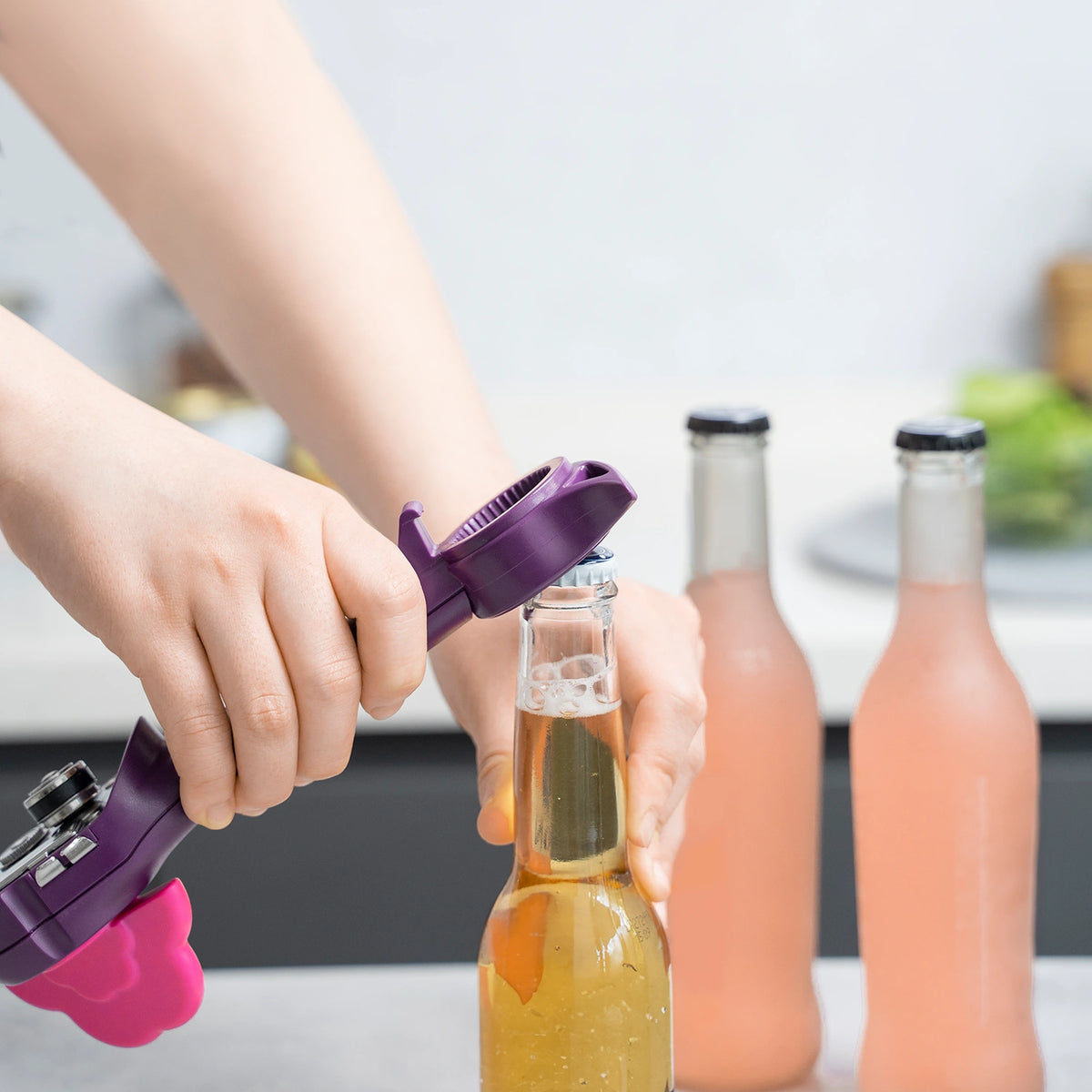 Eine Hand öffnet mit einem Flaschenöffner der Firma Kochblume GmbH eine Glasflasche, im Hintergrund sind zwei rosa Getränke zu sehen.