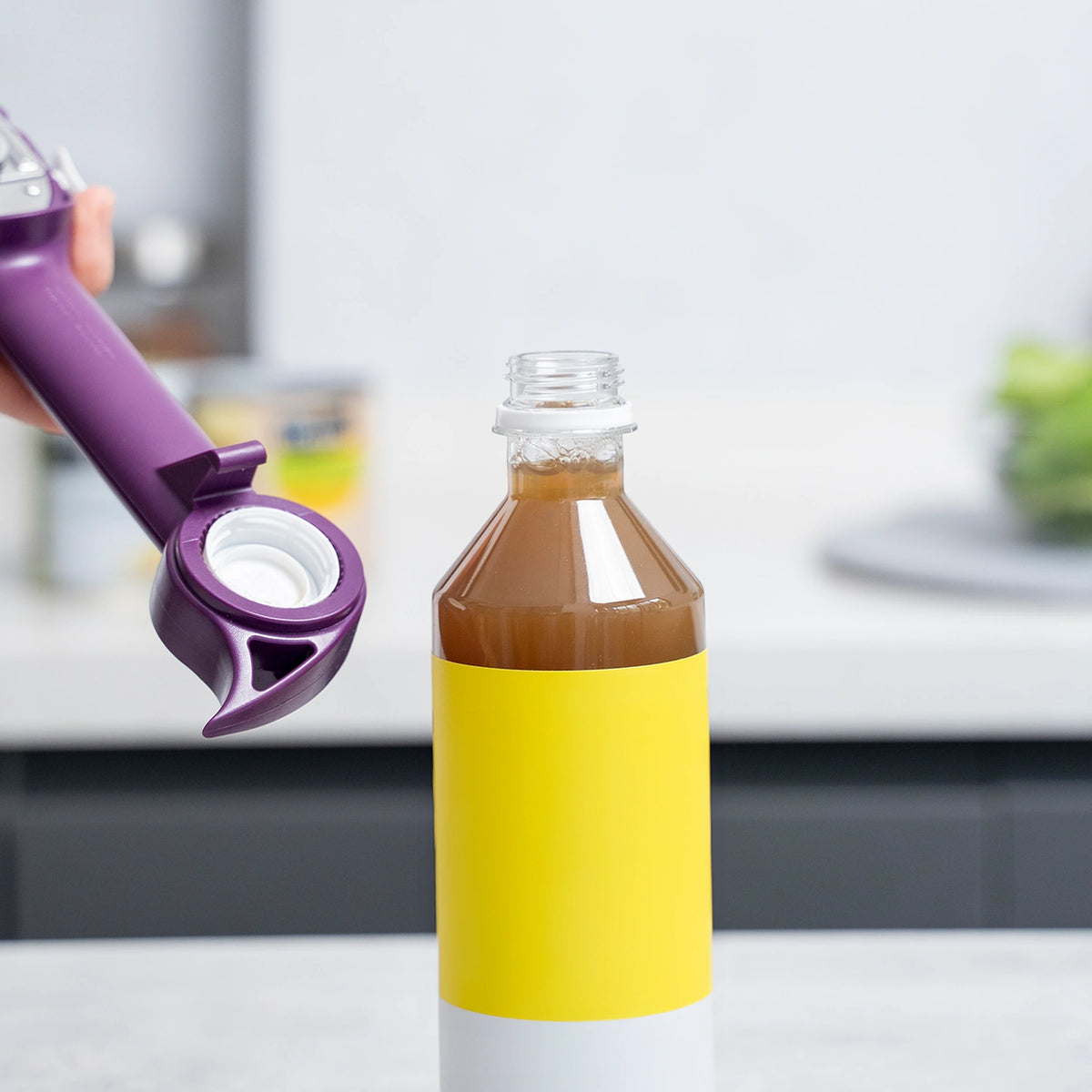 Ein Dosenöffner der Kochblume GmbH öffnet auf einer Küchenarbeitsplatte eine Flasche, die mit einer braunen Flüssigkeit gefüllt ist.