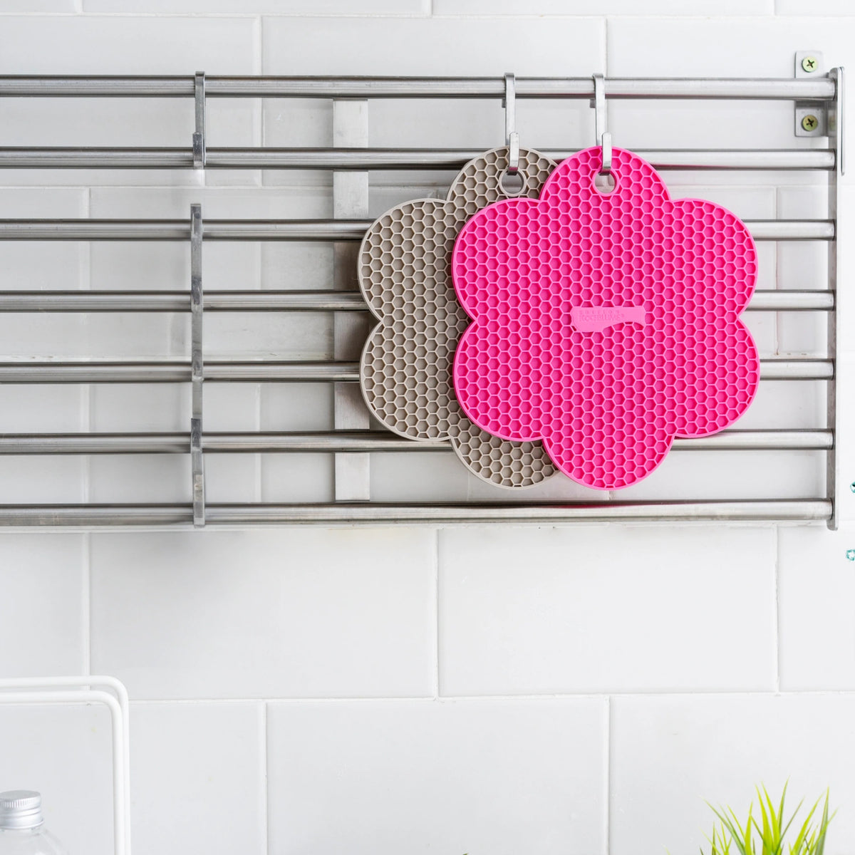 Zwei blumenförmige Silikon-Topfuntersetzer der Firma Kochblume GmbH – einer in Rosa und einer in Grau – hängen an einem Metallgestell vor einer weißen Fliesenwand. Ihre verspielte Anordnung und präzise Platzierung erinnern an ein Bild-Sprite, das an sorgfältig gestaltete Elemente erinnert, die mit CSS-Rahmentechniken hervorgehoben wurden.