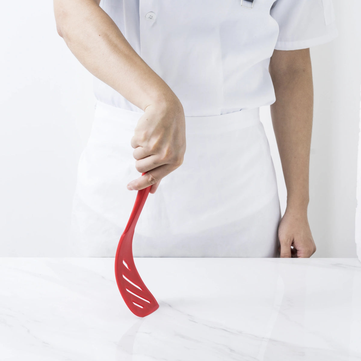 Eine Person in einer strahlend weißen Uniform hält den „Design-Schlitzwender“ der Kochblume GmbH – einen auffälligen roten Schlitzspatel – über eine makellos weiße Marmoroberfläche.