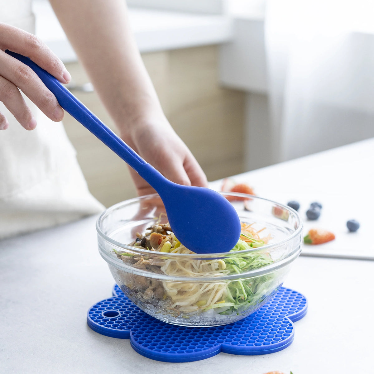 A person gently mixes noodles and fresh vegetables in a glass bowl using the Kochlöffel from Kochblume GmbH, atop a blue mat in a cozy kitchen setting.