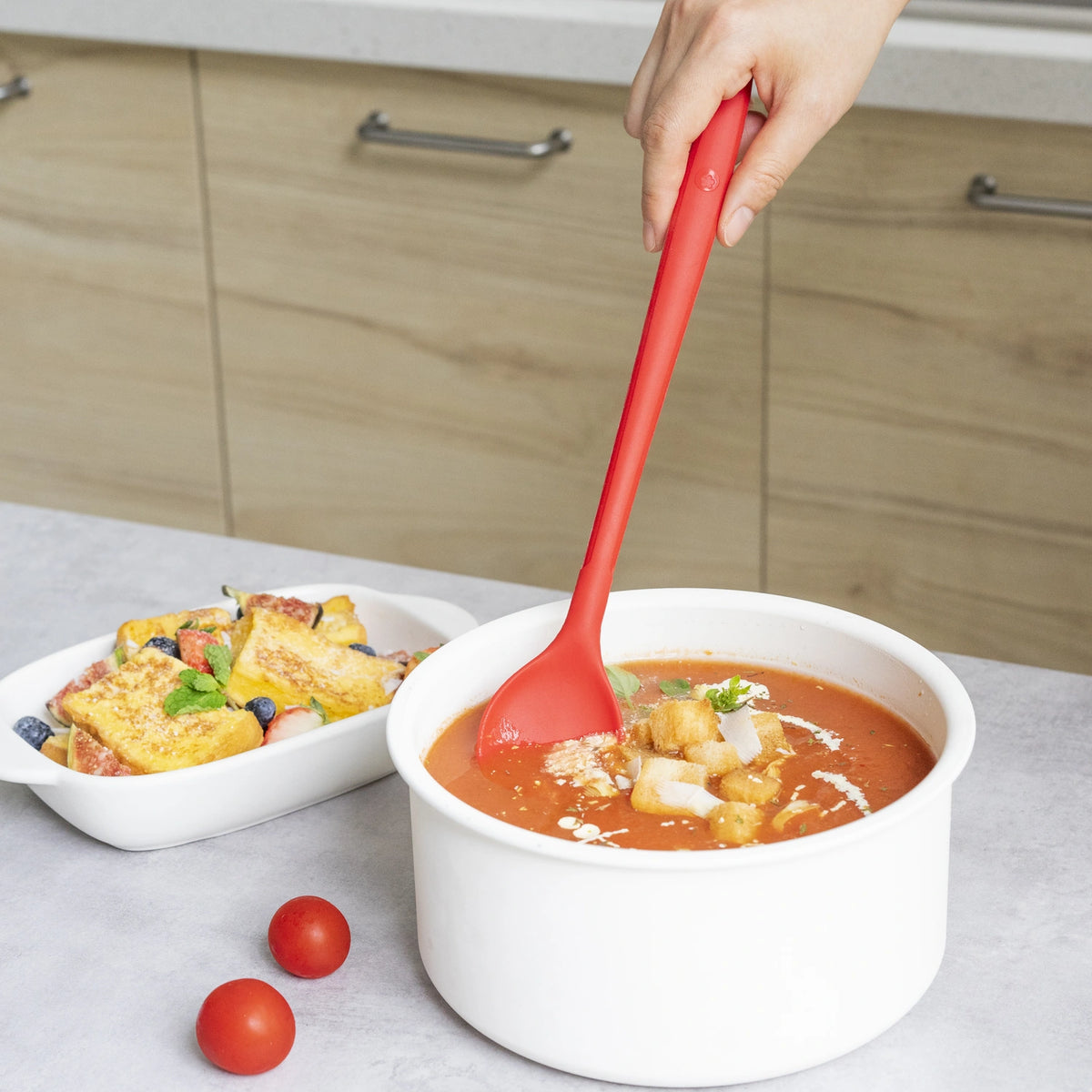 Stirring tomato soup in a white pot with the Kochlöffel by Kochblume GmbH, surrounded by a vibrant fruit salad and fresh tomatoes on the counter.