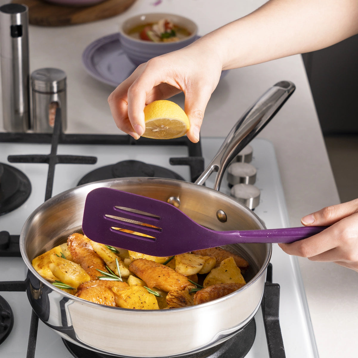 Eine Hand presst eine frische Zitrone über gewürzte Kartoffeln, die in einer Pfanne auf dem Herd garen, und rührt die Zutaten dabei vorsichtig mit dem leuchtend violetten Flexwender der Kochblume GmbH um.