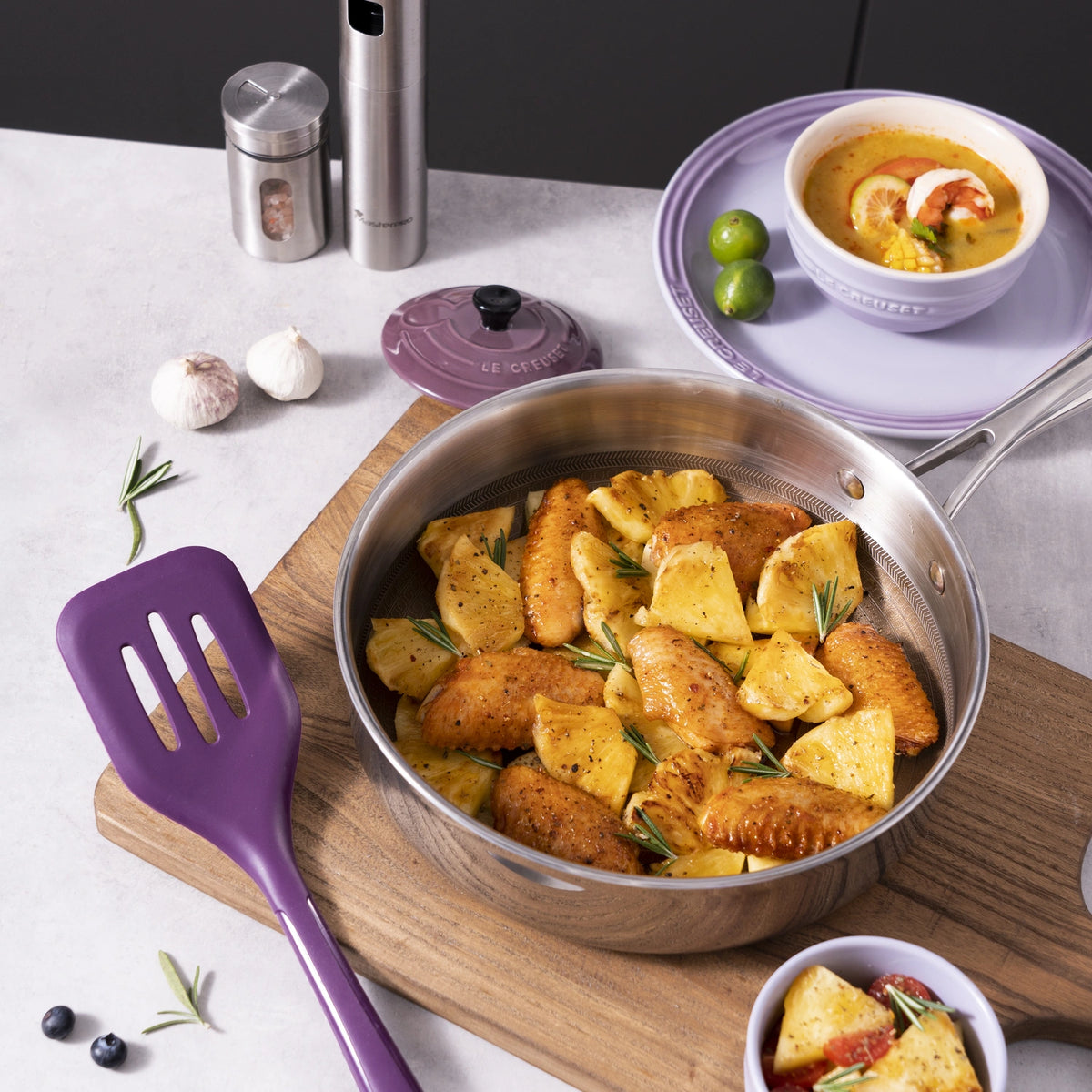 Neben einer Schüssel mit Curry und Küchenutensilien steht eine Pfanne mit gegartem Hähnchenfleisch und Ananas – alle mit dem Flexwender der Kochblume GmbH –, wodurch eine lebendige und funktionale Küchenszene entsteht.