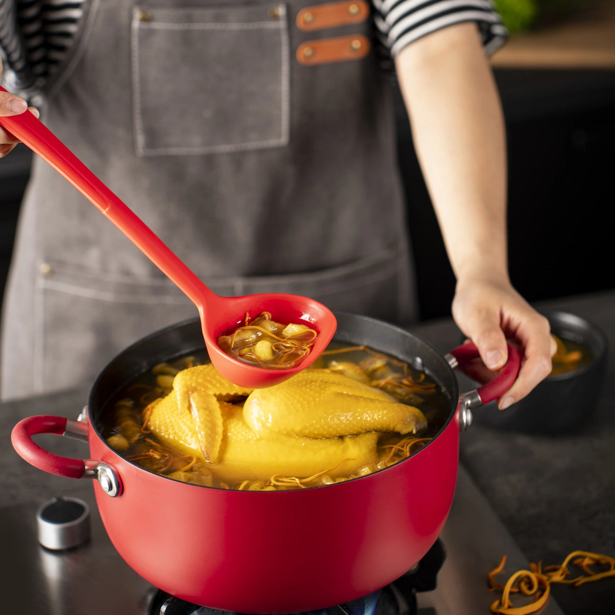 Eine Person schöpft mit der Schöpfkelle der Firma Kochblume GmbH Brühe aus einem roten Topf mit Hühnersuppe, in der ein ganzes Huhn und frische Kräuter in der dampfenden Suppe zu sehen sind.