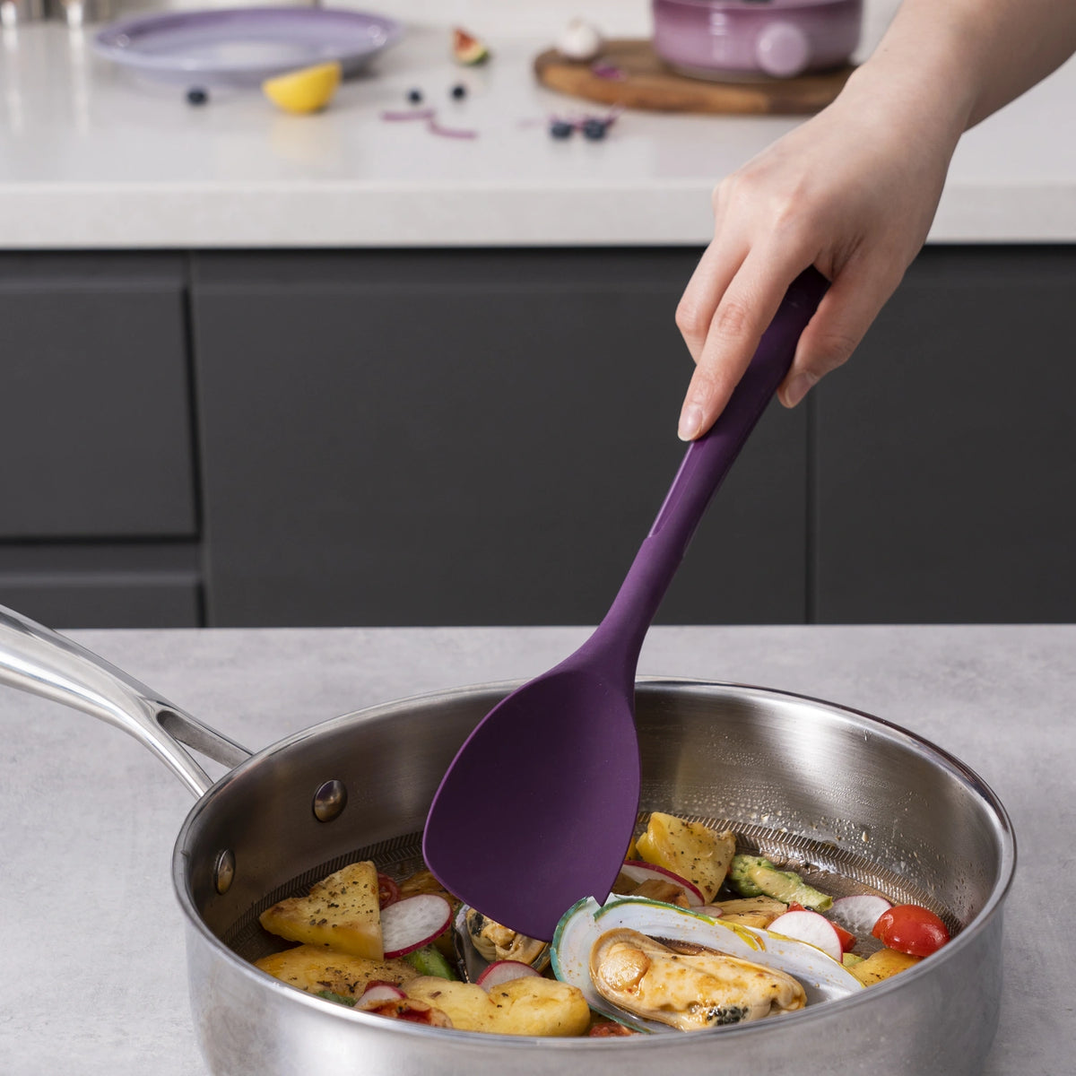 Gemüse wird von Hand in einer Edelstahlpfanne mit dem violetten „Flexlöffel“-Pfannenwender der Kochblume GmbH auf einer Küchenarbeitsplatte zubereitet; die bereitgestellte Produktbeschreibung enthält jedoch lediglich CSS-Code und keine Details zum „Flexlöffel“ selbst.