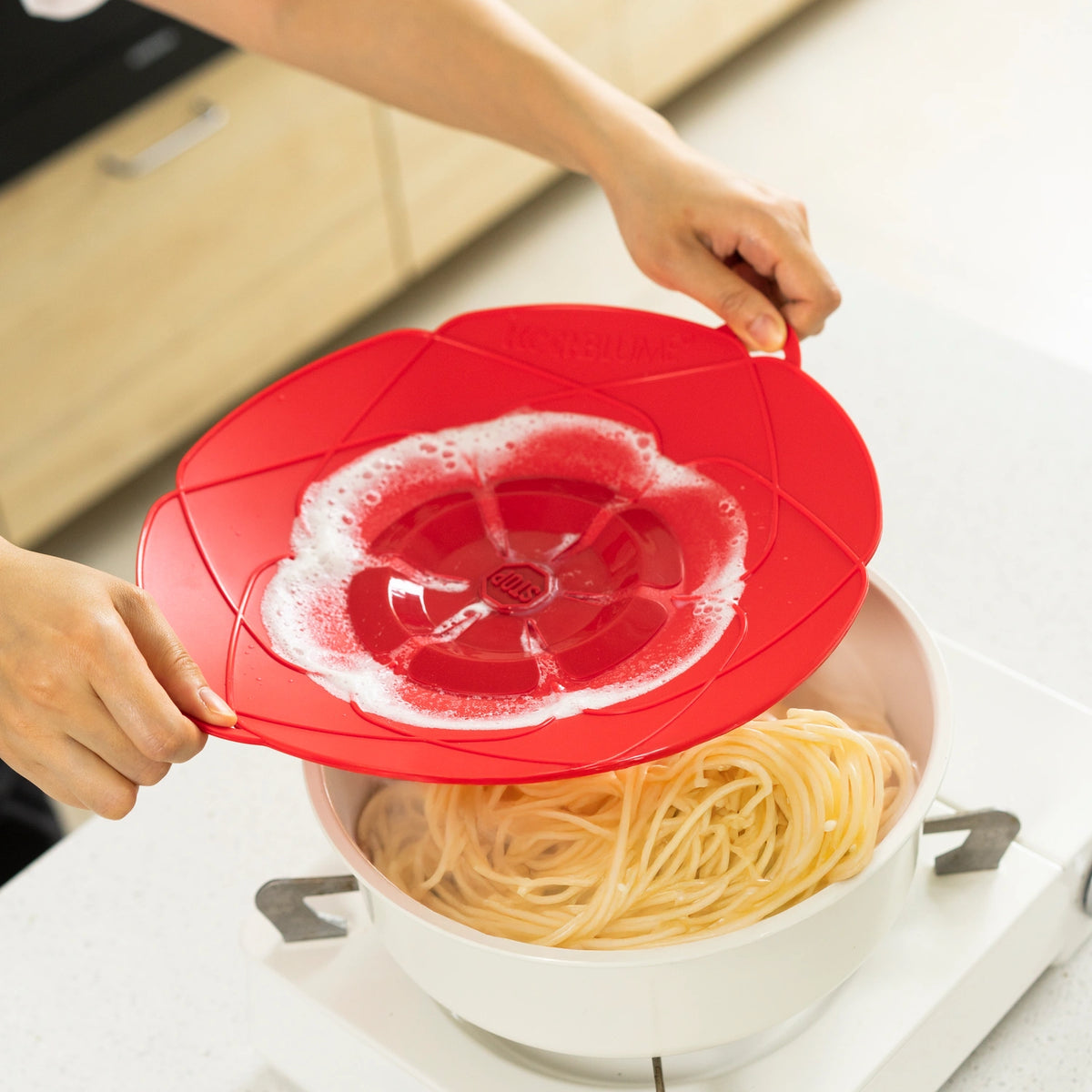 Eine Person nimmt den „Kochblume Überkochschutz“-Deckel der Firma Kochblume GmbH von einem Topf mit gekochten Spaghetti auf dem Herd ab.