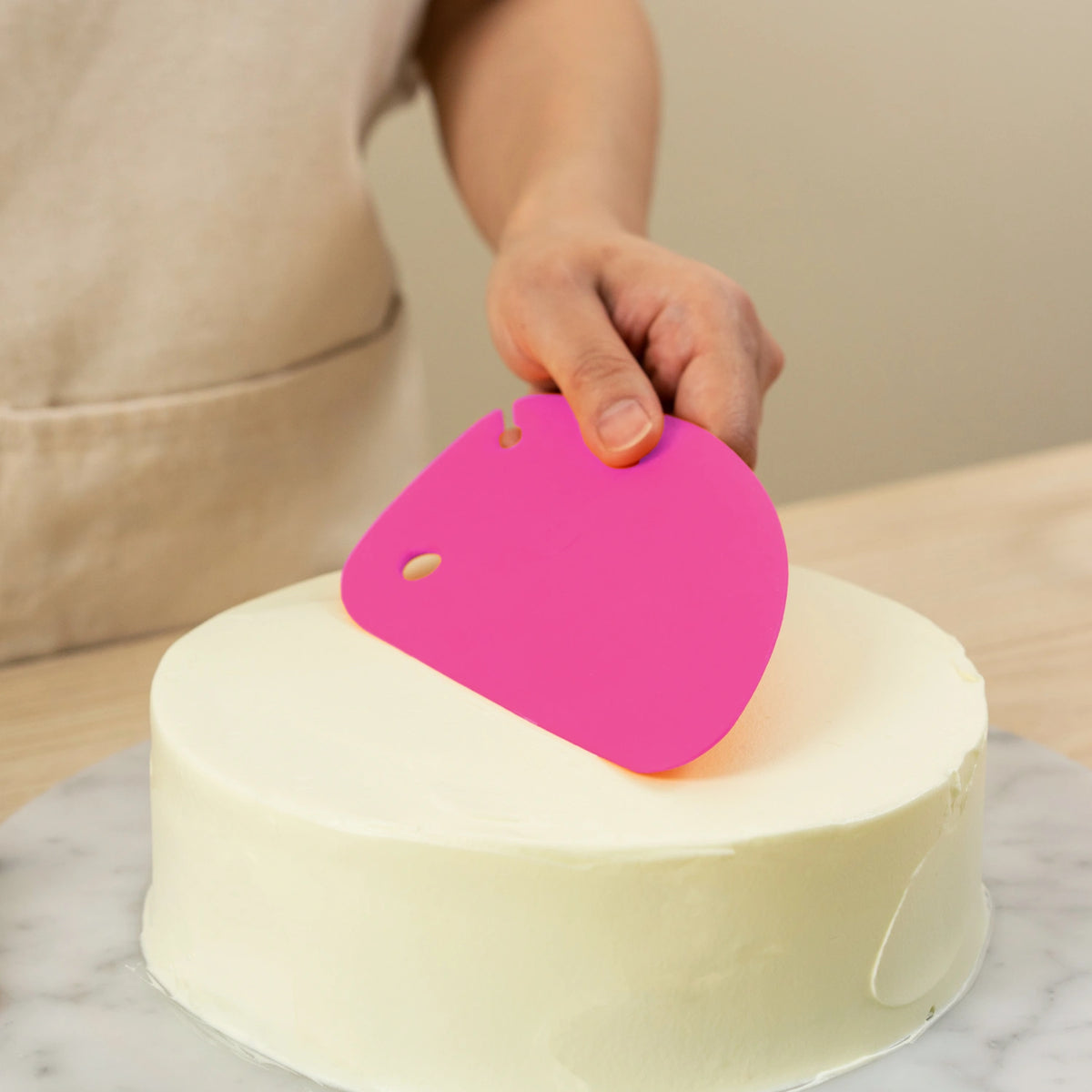 Eine Hand streicht mit der leuchtend pinkfarbenen Teigkarte der Kochblume GmbH auf einer Marmorplatte weiße Glasur auf einen Kuchen und sorgt so für makellose Ergebnisse bei Ihrem nächsten Backprojekt.