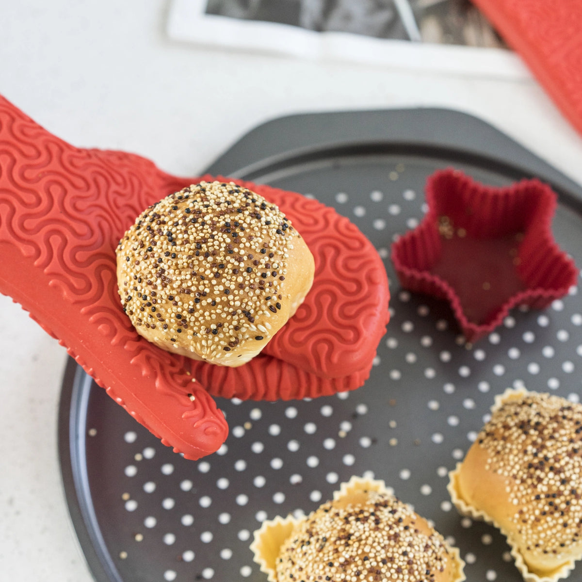 Ein roter Ofenhandschuh der Firma Kochblume GmbH hält ein mit Streuseln bestreutes Brötchen über einem Backblech, auf dem sich weitere Brötchen und eine sternförmige Backform befinden; die bereitgestellte Produktbeschreibung enthält jedoch lediglich CSS-Code und keine tatsächlichen Produktinformationen.