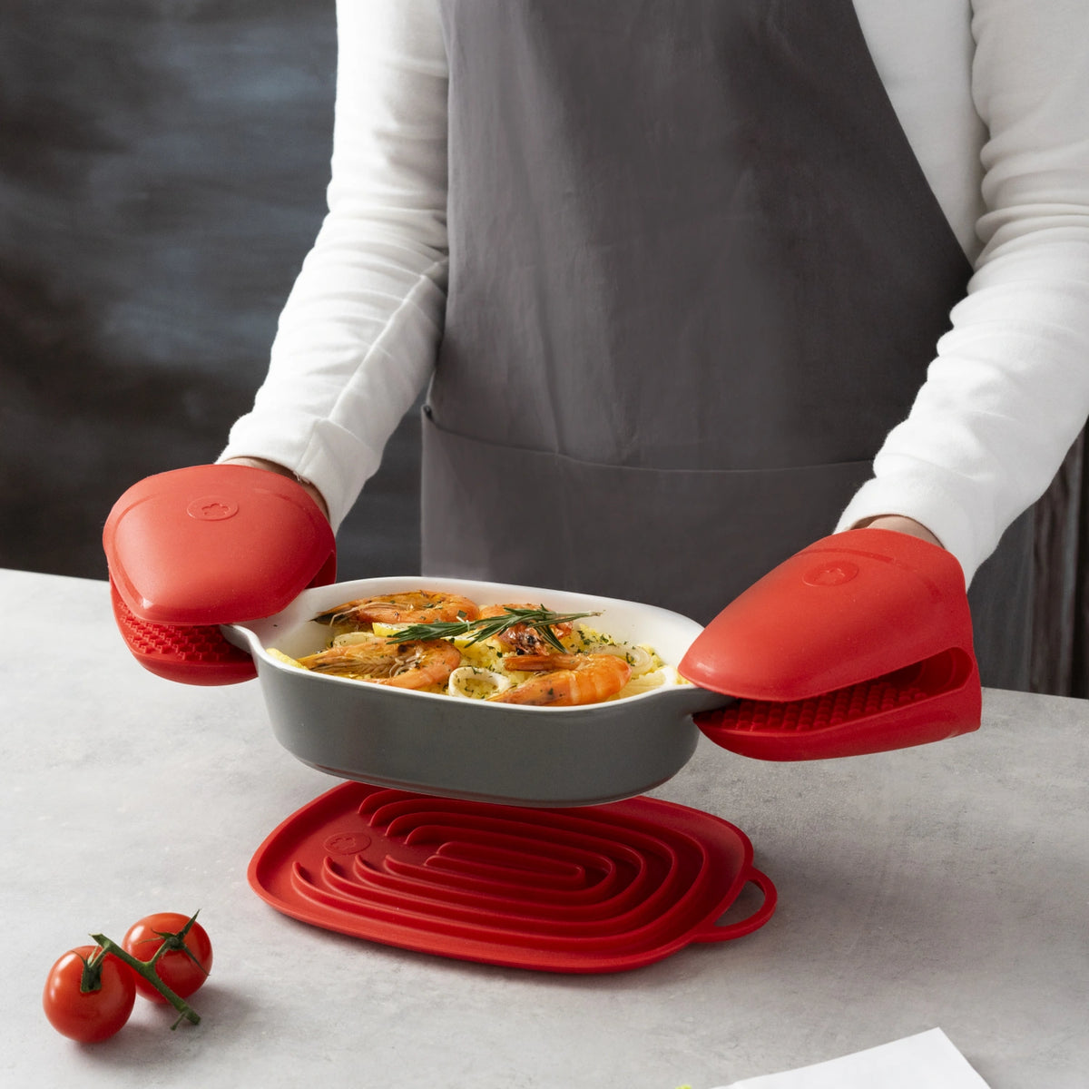 Eine Person, die die roten Ofenhandschuhe „Thermo-Fingerschutz“ der Kochblume GmbH trägt, hält eine Auflaufform in der Hand; auf dem Tisch stehen Tomaten und ein roter Untersetzer.