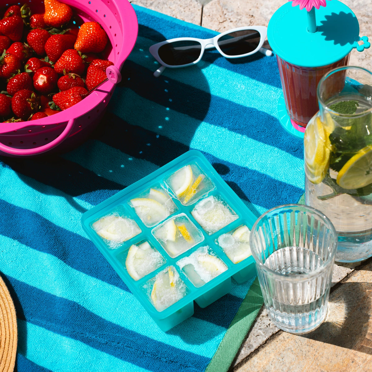 Eiswürfel aus der Eiswürfelform der Kochblume GmbH mit frischen Zitronenscheiben, einem Glas Wasser, Erdbeeren und einer Sonnenbrille liegen auf einem gestreiften Handtuch im Freien – perfekt zum Entspannen in der Sonne.