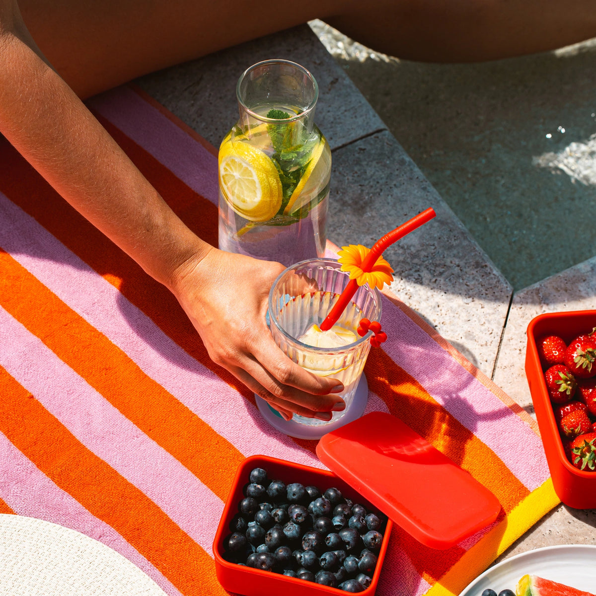 Eine Hand hält einen Trinkhalm der Kochblume GmbH in einem Glas am Pool, neben frischem Obst und einem Wasserkrug auf einem gestreiften Handtuch.