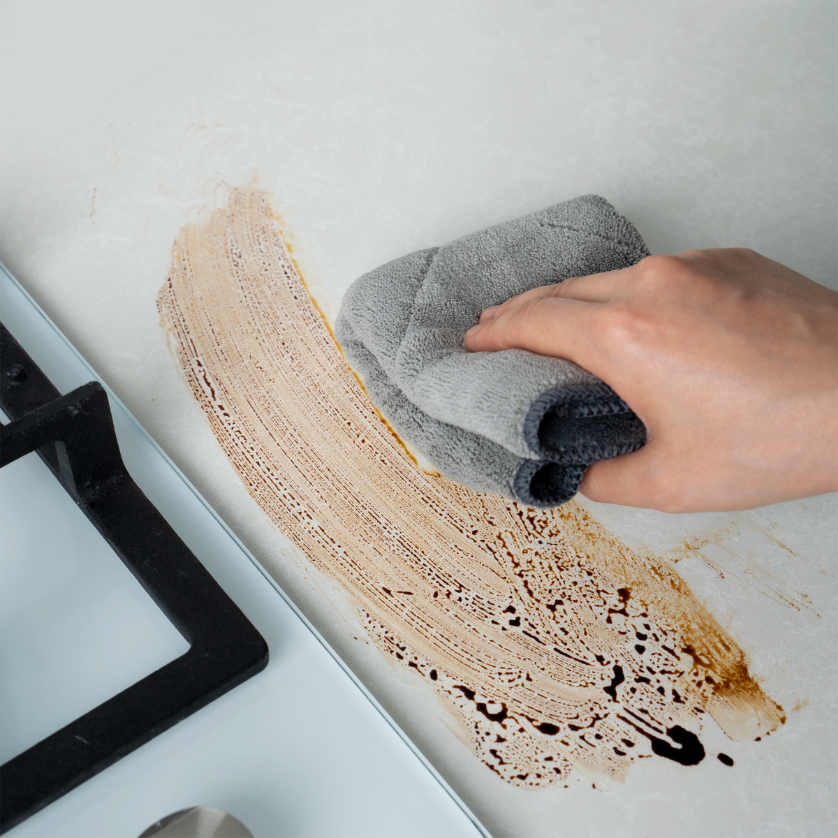 Mit einem grauen Tuch wird ein Kochfeld von Hand abgewischt, um eine braune Flüssigkeit von der weißen Oberfläche zu entfernen – unter Verwendung des Mikrofasertuchs der Kochblume GmbH. Dieses hochwertige Mikrofasertuch macht die Reinigung von Küchenoberflächen einfach und effizient und entfernt mühelos Verschüttetes, Fett und Flecken für ein makelloses Ergebnis.