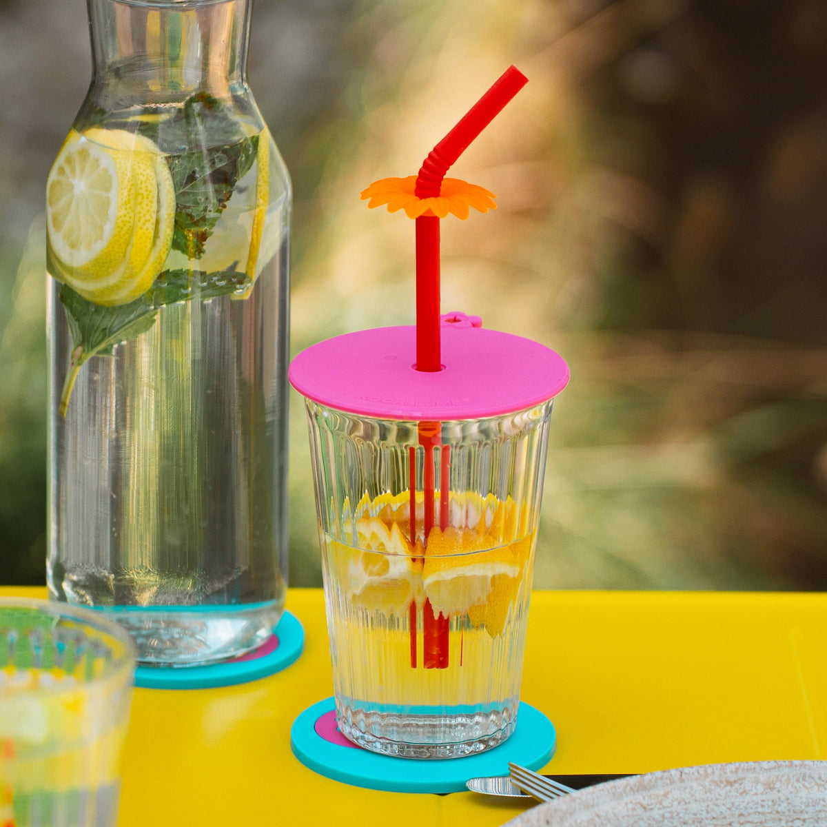 Ein Glas Zitronenwasser mit der „Glasabdeckung 3tlg.“ der Kochblume GmbH in Rosa als Deckel, dazu ein roter Strohhalm neben einer Karaffe mit Zitronenwasser – alles auf einem leuchtend gelben Tisch arrangiert und im frischen RTE-Stil mit klaren CSS-Rahmenlinien eingerahmt.