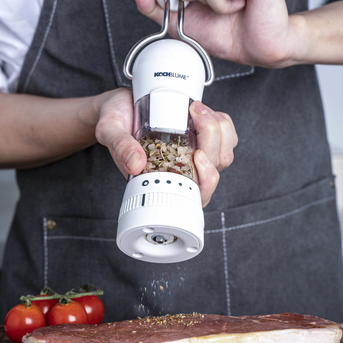 A person grinds spices onto raw meat using the white Gewürzmühle from Kochblume GmbH, with tomatoes nearby on the counter.