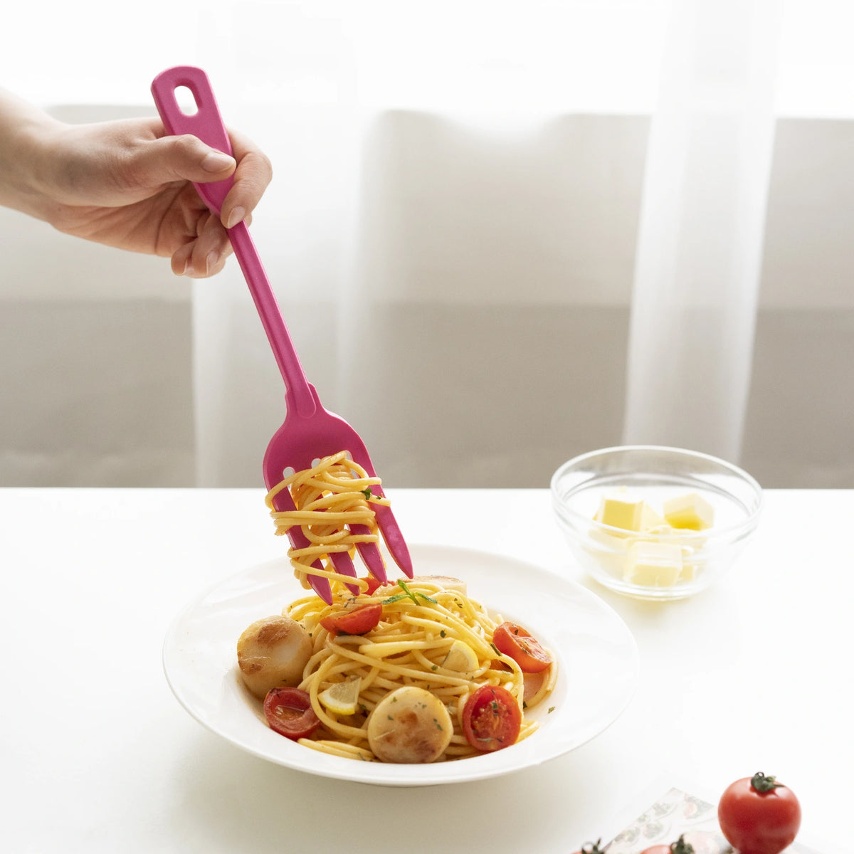 Mit der Küchengabel der Firma Kochblume GmbH werden Nudeln über einem Teller Spaghetti mit Tomaten und Jakobsmuscheln von Hand aufgerollt; die bereitgestellte Produktbeschreibung enthält jedoch lediglich CSS-Styling-Code und keine beschreibenden Informationen zu dem Küchenutensil.