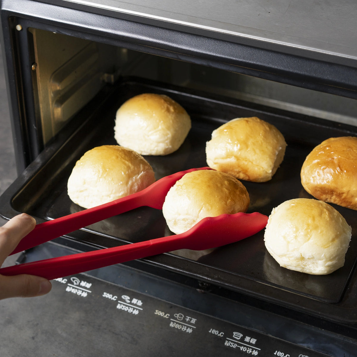 Eine Hand hebt mit der roten Gourmetzange der Kochblume GmbH vorsichtig ein goldbraunes Brötchen von einem Backblech im Ofen und demonstriert so die richtige Handhabungstechnik.