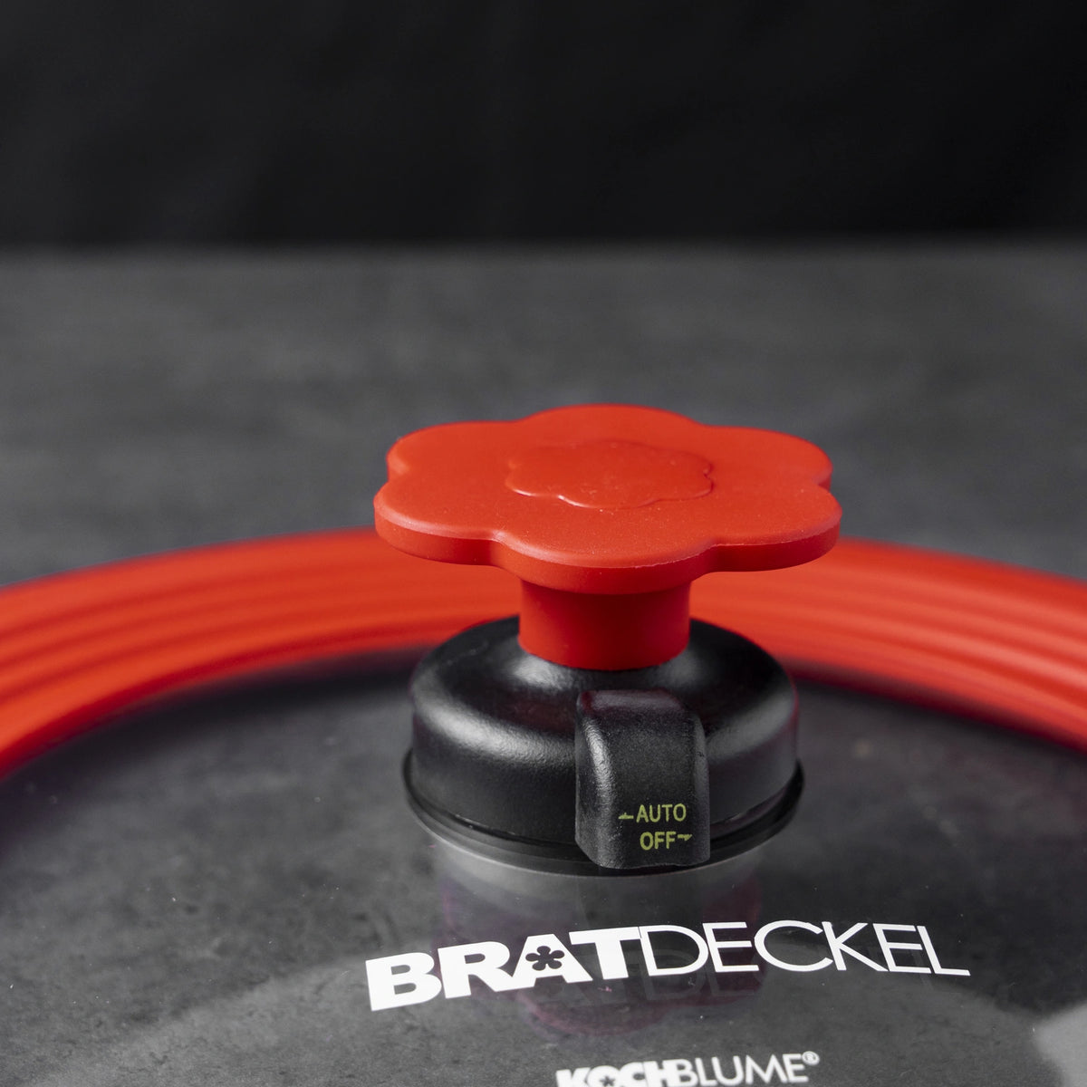 Den Glasdeckel „Bratdeckel“ der Firma Kochblume GmbH krönt ein roter, blumenförmiger Griff. Er verfügt über einen AUTO/OFF-Schalter und besticht durch schlanke, moderne Linien, die ihm eine zeitgemäße Ästhetik verleihen.