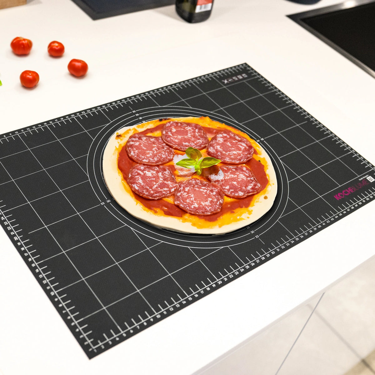 A pizza with sliced salami and fresh basil sits on a Kochblume GmbH Gewebematte on a white kitchen counter.