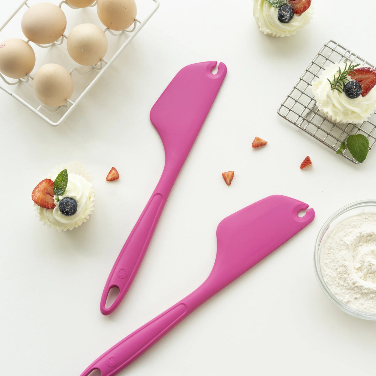 Two pink Allzweckschaber spatulas from Kochblume GmbH rest on a white surface, surrounded by eggs, cupcakes, flour, and scattered berries—setting the perfect scene for your next baking adventure. With these charming spatulas as your kitchen companions, homemade treats are just a step away.