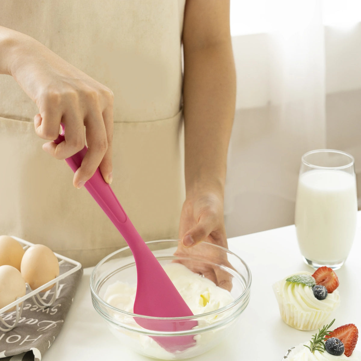 A person mixing batter in a glass bowl using the Allzweckschaber by Kochblume GmbH, featuring its signature pink color; eggs and cupcakes are nearby, with a glass of milk visible in the background.