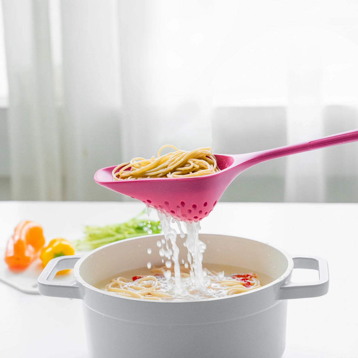 Ein rosafarbener Sieblöffel der Kochblume GmbH lässt Spaghetti über einem weißen Topf abtropfen, im Hintergrund sind verschwommene Gemüsestücke zu sehen – eine perfekte Ergänzung für Ihr praktisches Küchenzubehör.