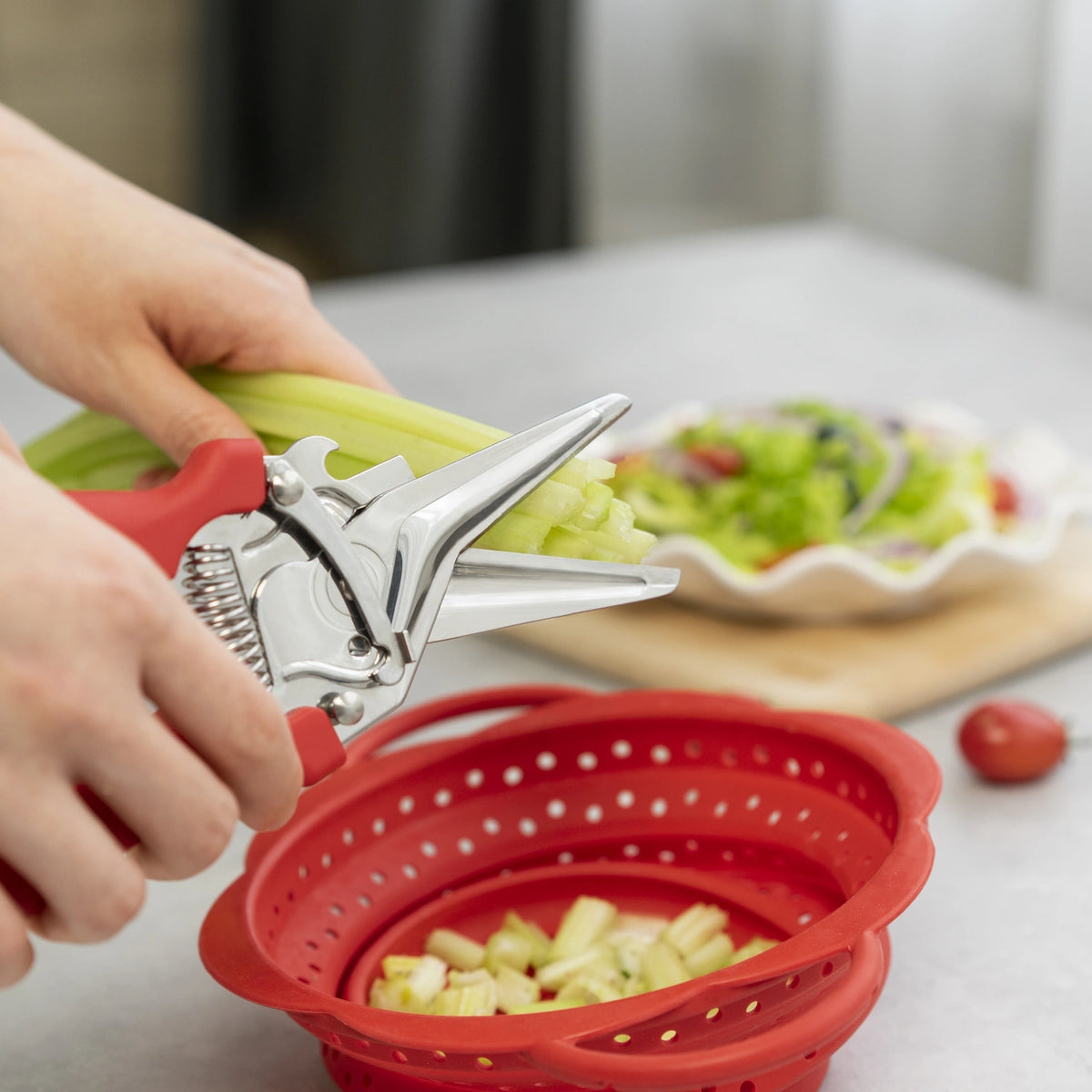 Hände, die mit der roten Allzweckschere der Kochblume GmbH Sellerie über einem roten Sieb schneiden, im Hintergrund steht ein frischer Salat auf der Arbeitsplatte.