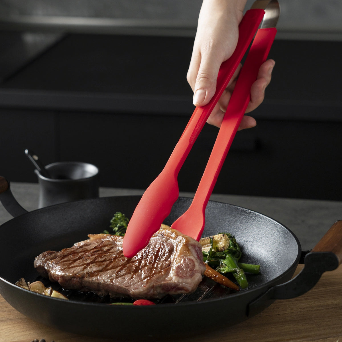 Hand wendet mit der „Gourmetzange“ der Kochblume GmbH ein Steak in einer Pfanne mit Gemüse auf einer Küchenarbeitsplatte.