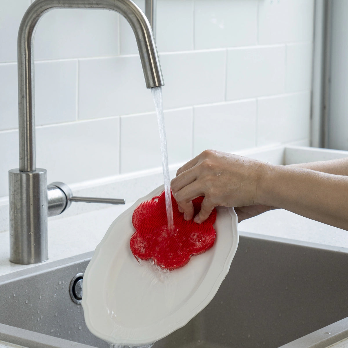 Eine Person spült einen weißen Teller unter dem Wasserhahn mit einem roten „Swisch“-Scheuerschwamm der Firma Kochblume GmbH in einer Küchenspüle.