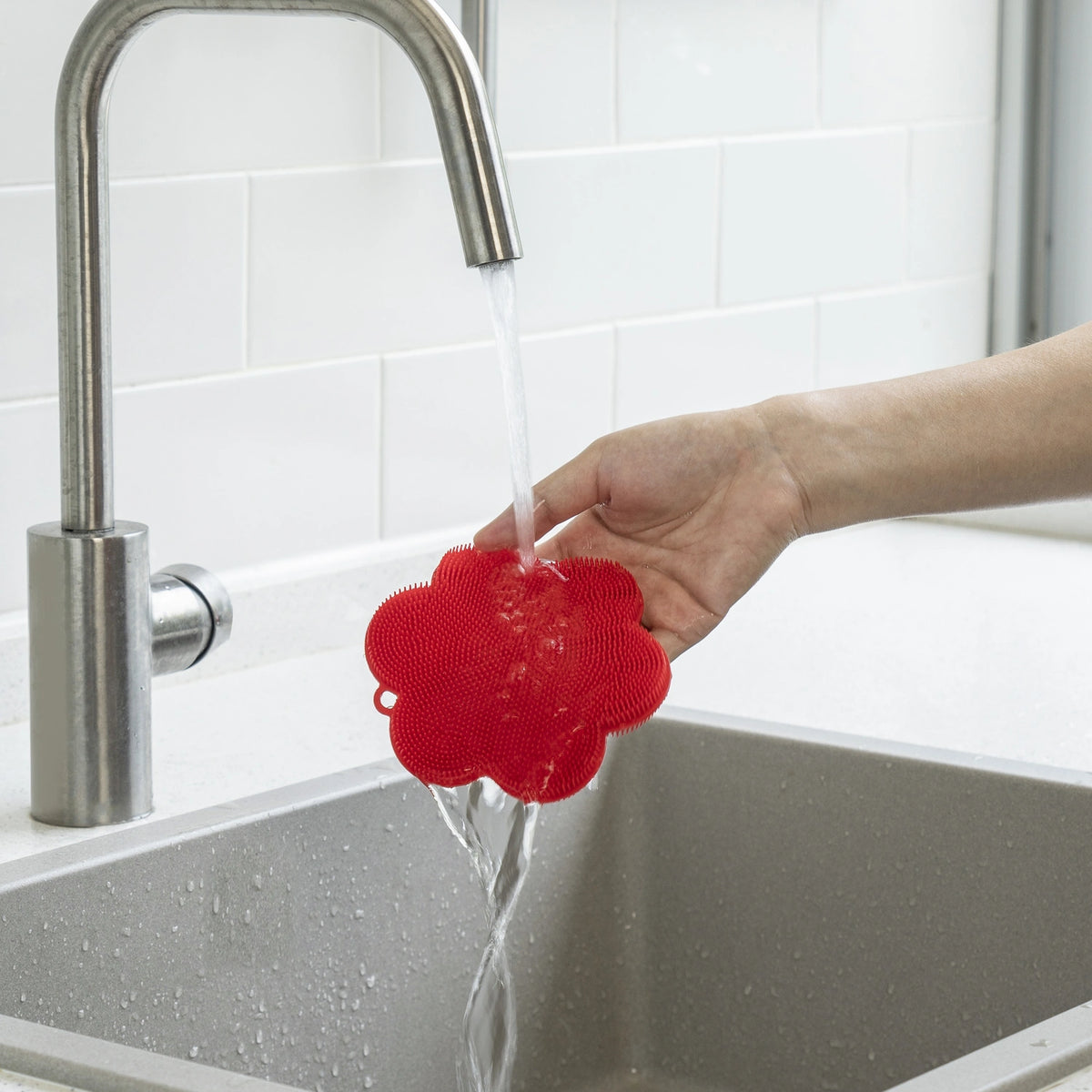 Eine Hand hält den „Swisch“ der Kochblume GmbH, einen roten Silikon-Scheuerschwamm, unter fließendes Wasser aus dem Küchenhahn über einem Spülbecken – und zeigt damit, wie einfach er zu reinigen und wie praktisch er für alltägliche Küchenarbeiten ist.