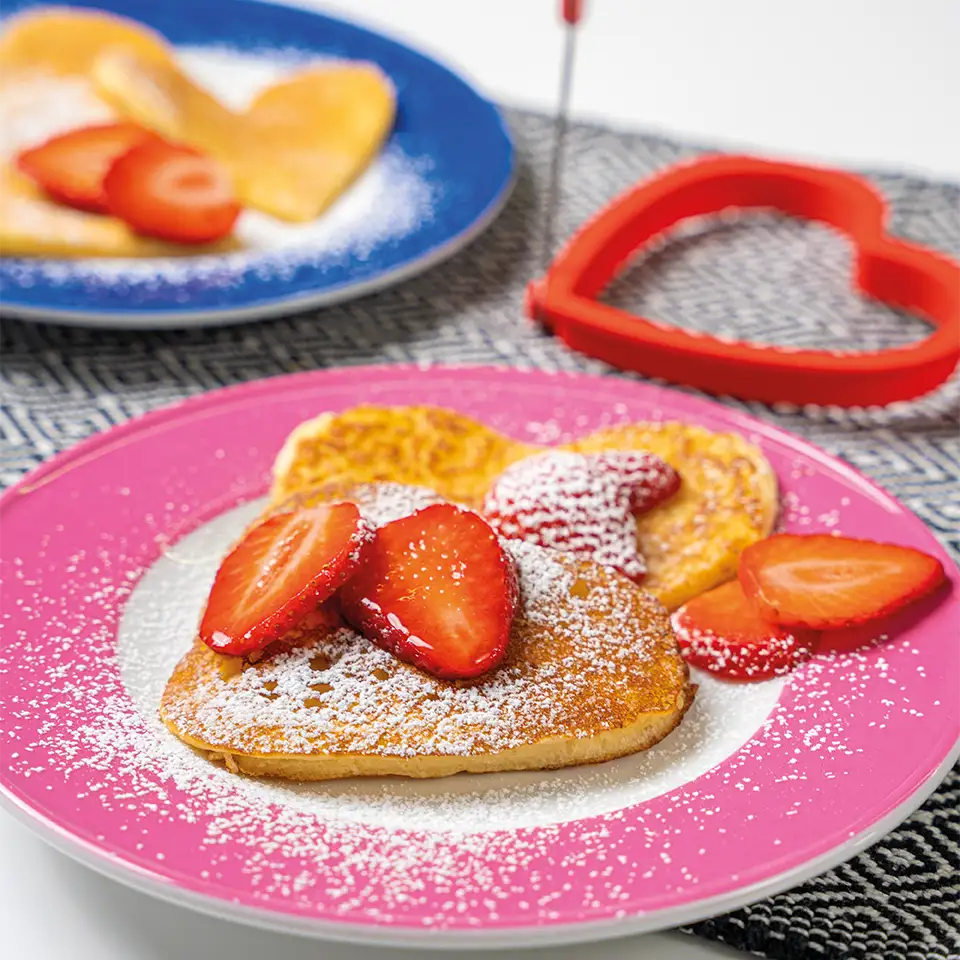 Herzförmige Pfannkuchen, gebacken mit den „Eierformen“ der Kochblume GmbH, garniert mit Erdbeerscheiben und Puderzucker auf einem rosa Teller – perfekt für kreative Frühstücksideen.