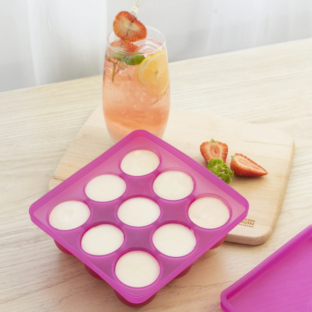 Eine Eiswürfelform der Kochblume GmbH aus rosa Silikon, mit der sich runde Eiswürfel herstellen lassen und die jedem Getränk eine verspielte Note verleiht, steht neben einem fruchtigen Drink, der mit Erdbeerscheiben garniert ist. Mit ihrem farbenfrohen Design und ihrer praktischen Anwendbarkeit eignet sich diese Form ideal für all Ihre gekühlten Getränke.