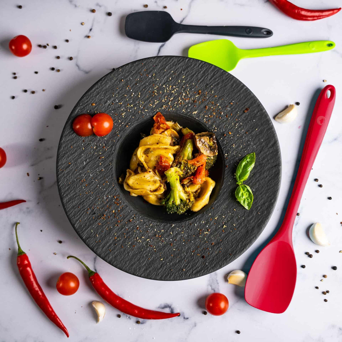 Nudeln mit Gemüse in einer schwarzen Schüssel, umgeben von Tomaten, Chilischoten, Knoblauch und bunten Pfannenwendern.