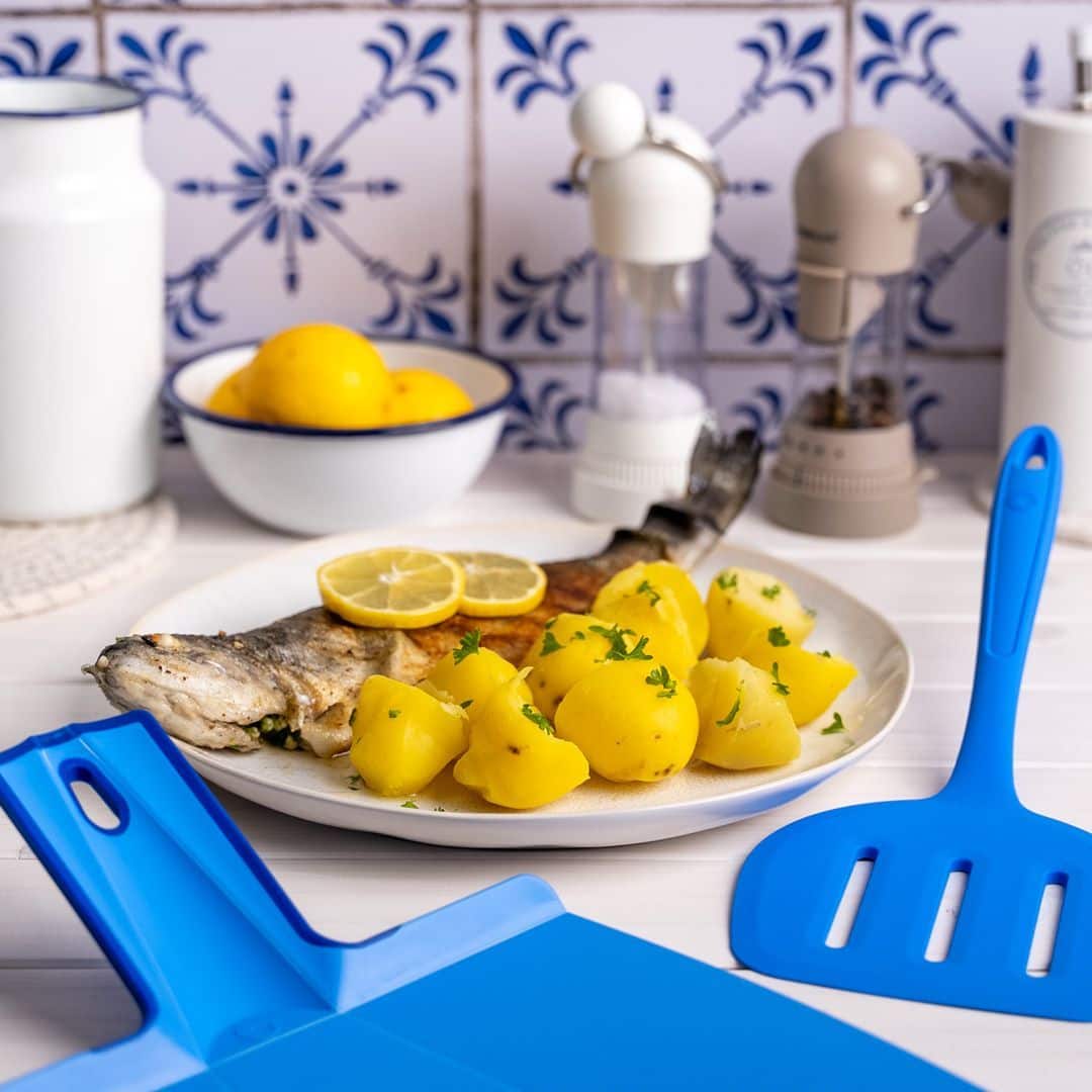 Gegrillter Fisch mit Zitrone und Kartoffeln auf einem Teller, blaues Besteck und im Hintergrund eine Schale mit Zitronen.