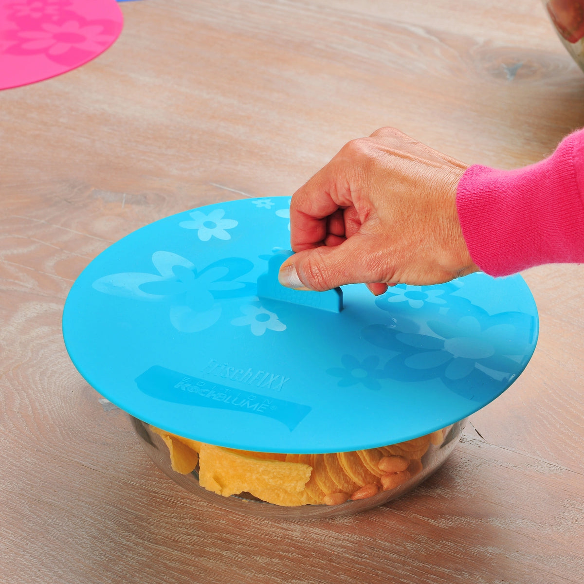 Eine Hand hebt einen blauen, mit Blumenmustern verzierten Frischfixx-Silikondeckel der Firma Kochblume GmbH von einer mit Chips und Snacks gefüllten Glasschüssel auf einem Holztisch.