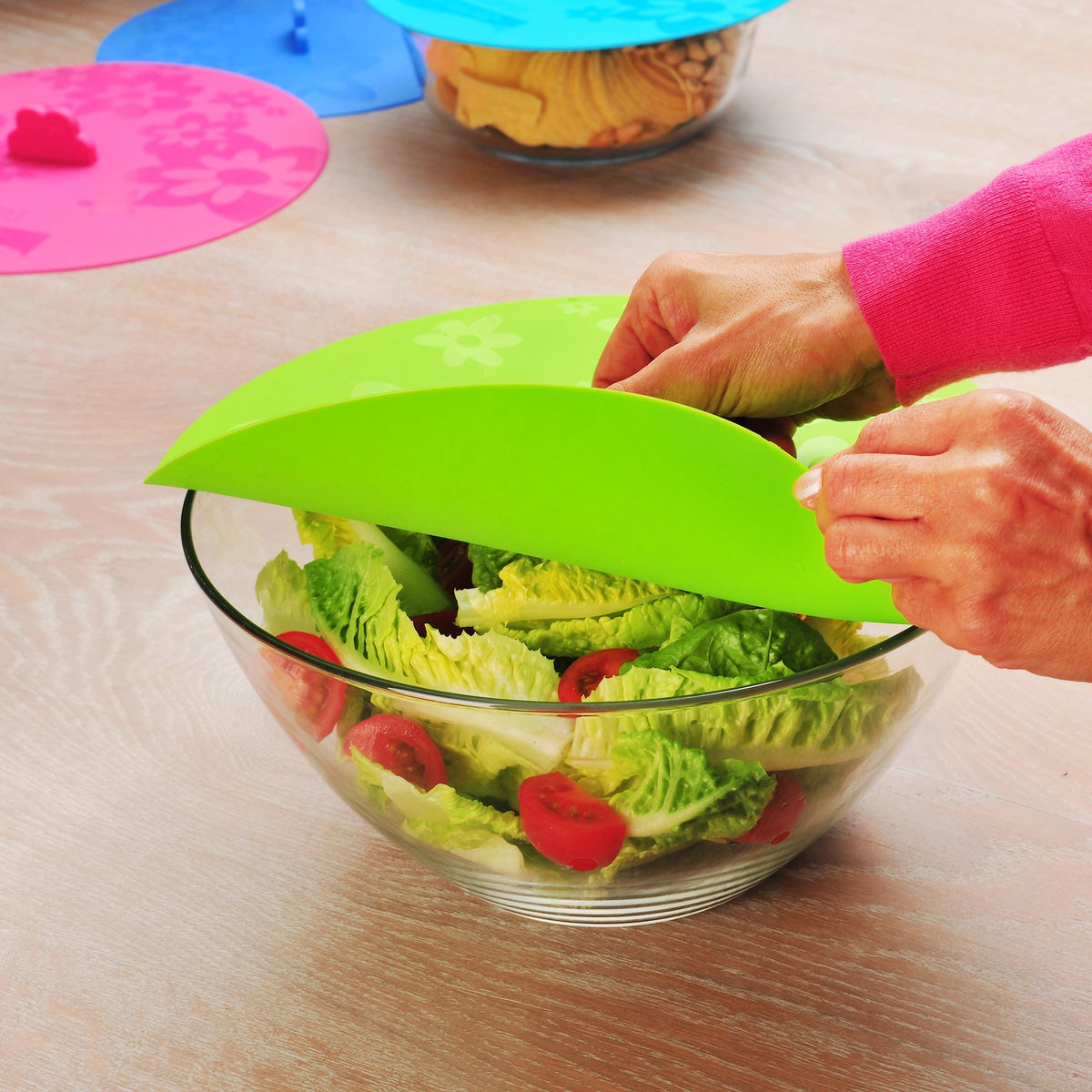 Hände decken eine Salatschüssel mit einem flexiblen grünen Frischfixx-Deckel der Kochblume GmbH ab und unterstreichen so den sicheren Sitz. Weitere Schüsseln im Hintergrund, die jeweils mit unterschiedlichen Frischfixx-Deckeln verschlossen sind, zeigen, wie verschiedene Behälter effizient frisch gehalten werden können.