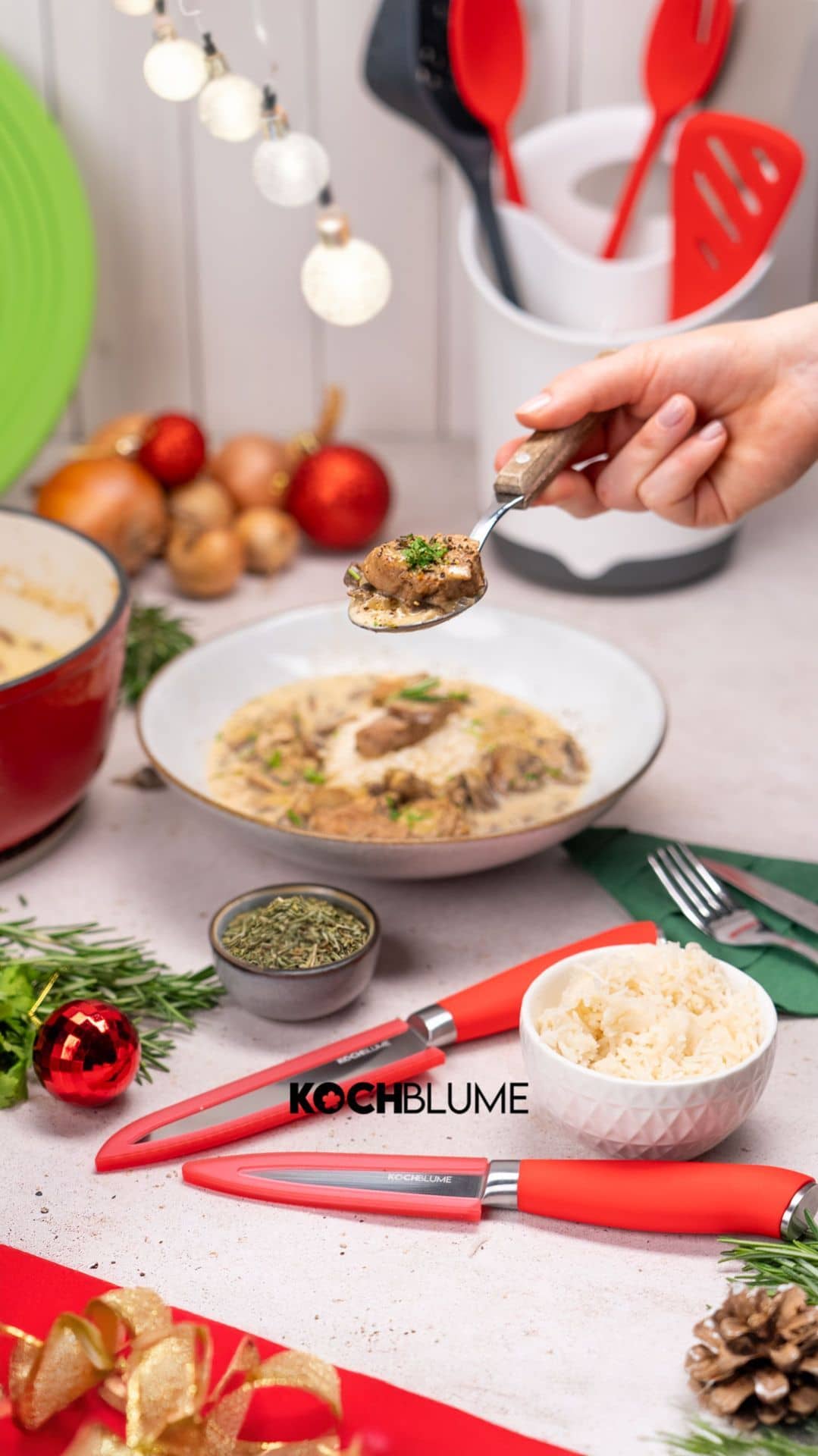 Eine Hand hält einen Löffel mit Essen über eine Schüssel; im Hintergrund sind Küchenutensilien und festliche Dekoration zu sehen.