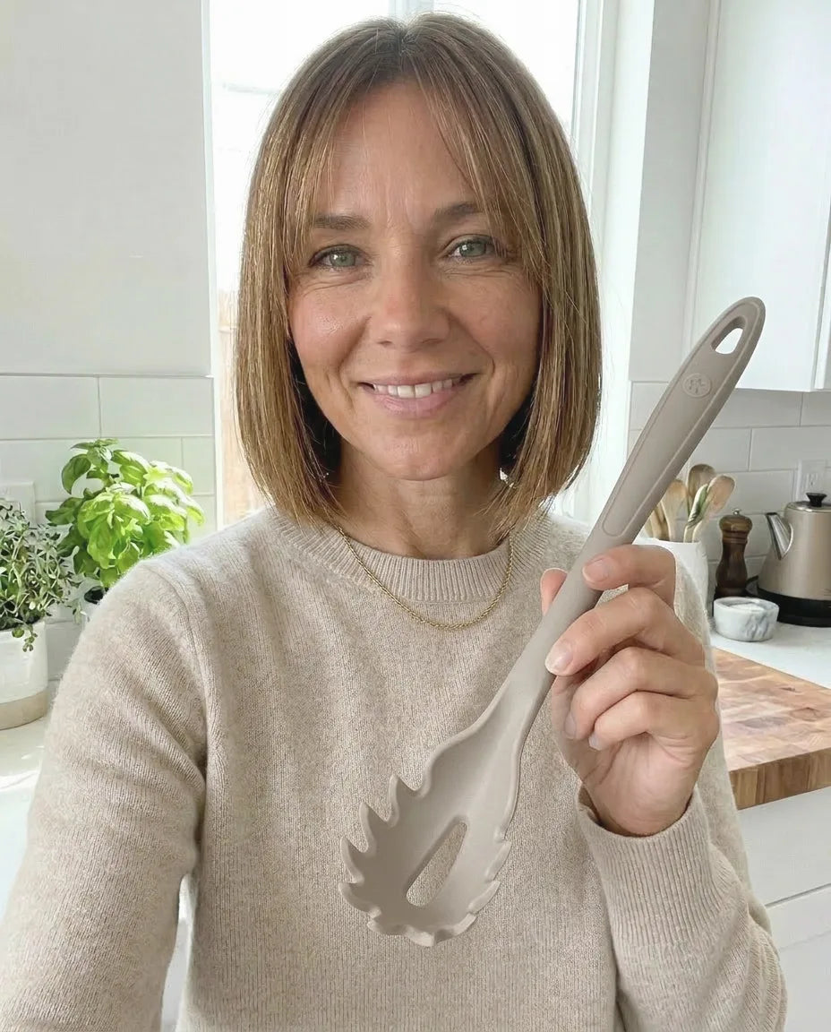Eine Frau lächelt in einer Küche und hält in ihrer rechten Hand einen beigen Nudelschaufel.
