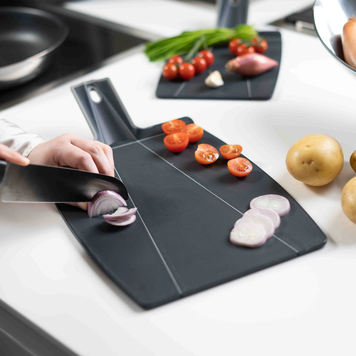 Eine Hand schneidet gekonnt eine rote Zwiebel auf dem „Faltschneidbrett“ der Kochblume GmbH, einem eleganten schwarzen Schneidebrett, während daneben frische Tomaten, Schalotten und Kartoffeln angeordnet sind – eine einladende Szene, die sich ideal für Produktbeschreibungen oder die Extraktion von Schlüsselwörtern eignet.